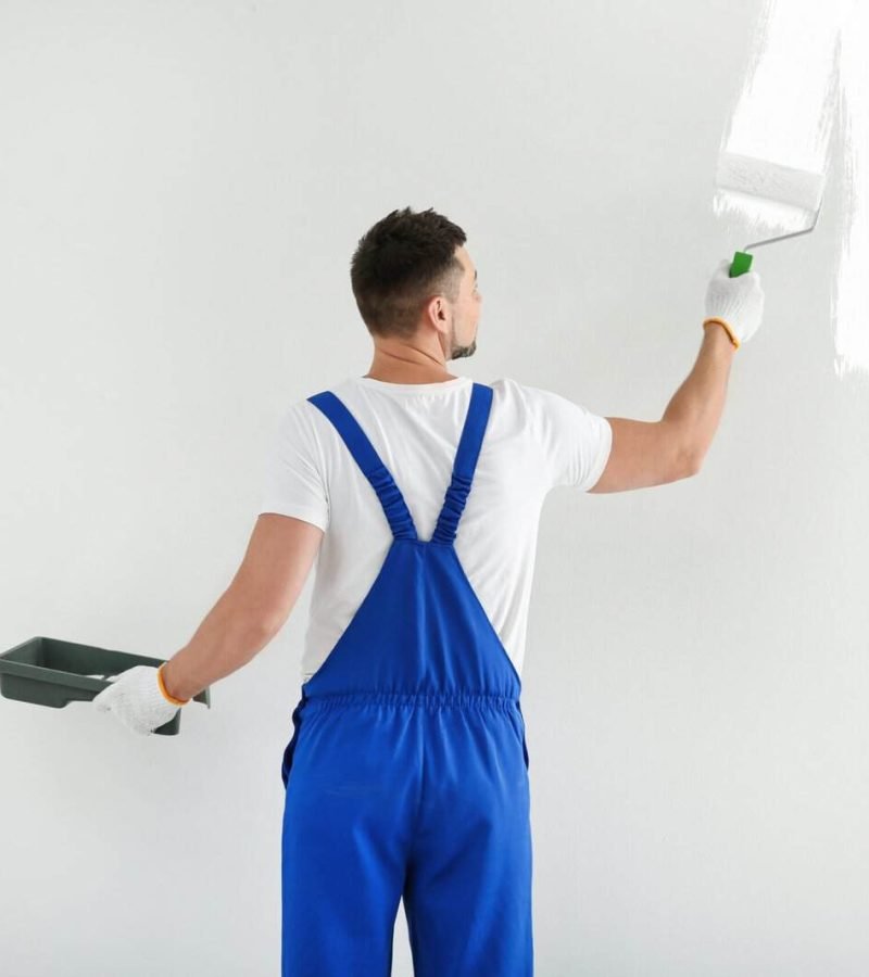 Painter in blue overalls painting a white wall with a roller brush.