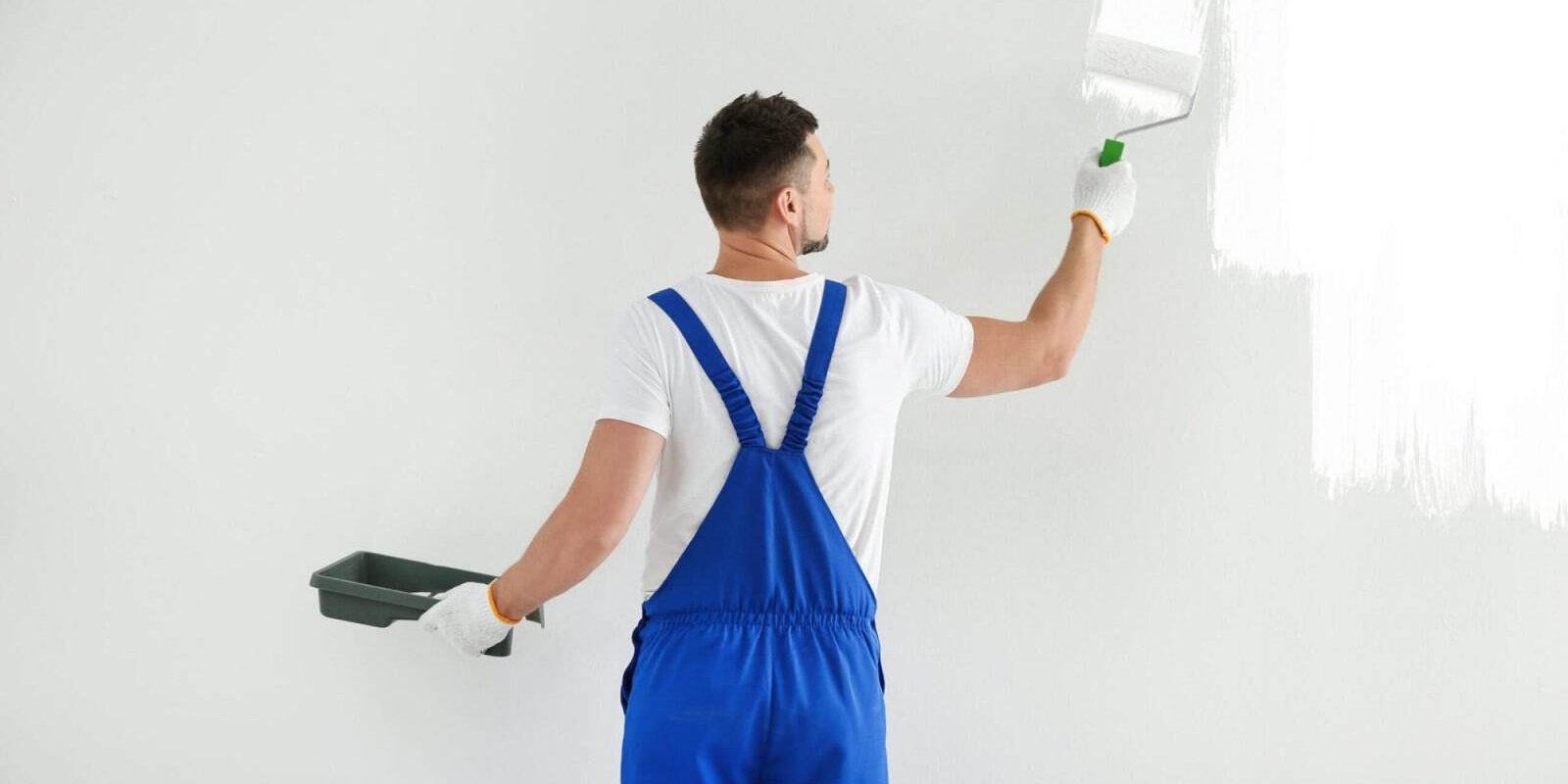 Painter in blue overalls painting a white wall with a roller brush.
