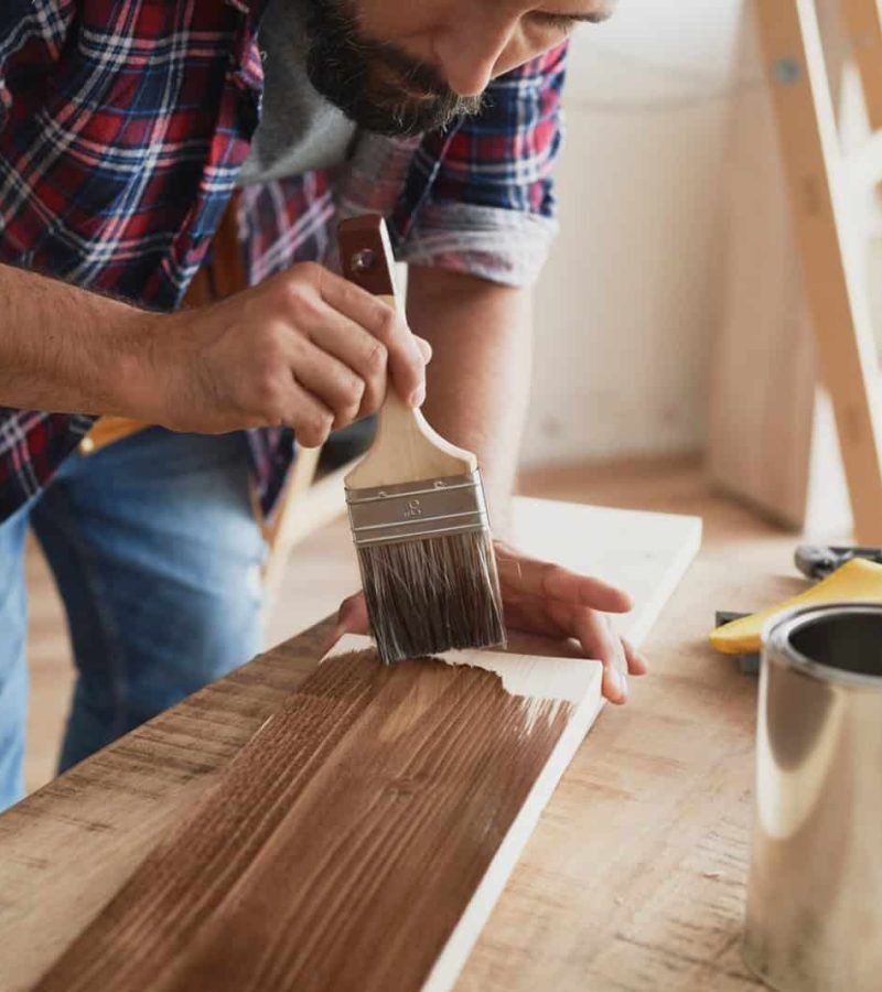 Man applying brown paint to wood surface with brush in workshop.