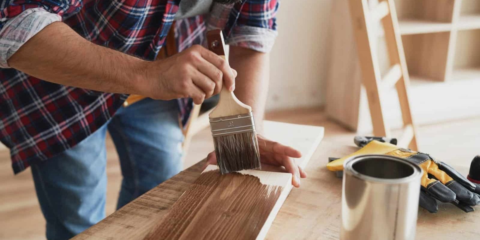 Man applying brown paint to wood surface with brush in workshop.